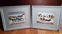 Two framed watercolor prints depicting early Americana folk scenes, displayed side by side on a wooden floor