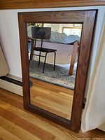 Front view of the vintage wall mirror propped against a baseboard heater and wooden floor. Shows the full reflective glass and wooden frame with natural finish.