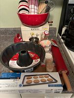 Photo showing the red mixing bowl, apple peeler, Betty Crocker nonstick Bundt cake pan, wooden rolling pin, box of silicone baking liners, whisk, oven mitts, and small baking accessories on kitchen counter.