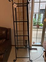 Full view of 5 tier black metal shelving unit with glass shelves and scroll design on top and feet positioned near window next to couch.