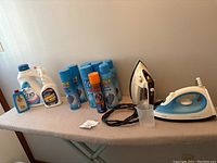 The full tabletop view showing vintage ironing board with cover, Philips iron, Black & Decker iron, multiple cans of spray starch and laundry detergent bottles, along with a small plastic cup.