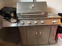 Full front view of the stainless steel KitchenAid gas barbecue grill showing five control knobs, side burner on left with cover, and double-door cabinet below.