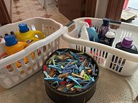 Overview of the three white plastic laundry baskets: two holding various opened laundry products and detergents, one empty. Large black container filled with multi-colored clothespins.