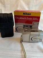 Display of Nikon Lite-Touch Zoom 110s film camera, original box, and black leather carrying case arranged on fabric surface