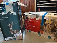 Two boxes of lighted animated grapevine standing deer shown in storage with worn tape and some damage to the boxes.