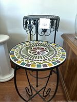 Front view of two metal plant stands, one taller with decorative scrollwork and one shorter with mosaic tile top in a geometric star and border pattern.