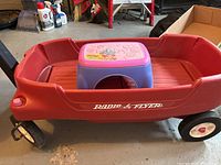 Photo of red Radio Flyer wagon showing one wheel and side with branding and safety belts inside, including the pull handle attachment and the purple step stool placed inside the wagon