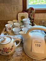 Overall view of tea set items including teapot, assorted teacups, saucers, mugs, and Sunbeam kettle on kitchen counter near window.