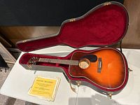Full view of Sovereign acoustic guitar in open black hard case with red velvet lining and some vintage sheet music placed beside it.