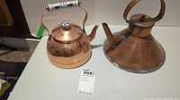 Photo of two copper kettles side by side, one polished with porcelain handle and knob, the other roughly shaped with patina.