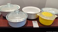 Five casserole bowls: four white CorningWare with glass lids and one yellow Pyrex bowl with sunflower lid and chip on rim, displayed on wooden surface.