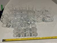 Wide view showing 21 various crystal glasses including stemmed glasses and tumblers arranged in rows with measuring tape showing scale.