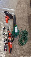 Full view of the Black & Decker lithium-ion weed trimmer with two batteries and a charger alongside the green electric leaf blower with attached extension cord coiled next to it on carpeted floor.