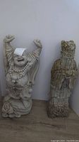 Front view of two concrete garden figurines against a white wall: Laughing Buddha with arms raised and an Asian-style figure with hands clasped.