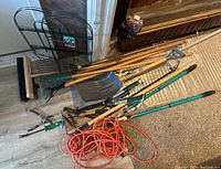 Full view of various garden tools laid on floor including shovels, rakes, pruning shears, and a folding wire fence panel leaning upright.