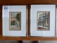Two watercolour paintings in wooden frames with white mats, showing building exterior scenes, one with arched windows and a tree and the other with balconies and detailed facade.