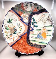 Full view of scalloped edge Japanese Imari charger plate with blue, orange, floral and figurative hand painted designs on surface displayed on black stand.