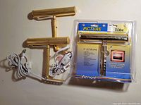 Two used brass finish picture lights with cords and in-line switches alongside one new light in sealed original Attron Picture Lite packaging.