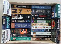 Overhead view of 21 VHS movie cases in a cardboard box showing classic movie titles.
