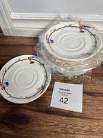 Stack of ten white bone china saucers with colorful floral pattern around edge, some individually wrapped.