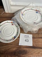 Set of ten bone china saucers stacked, some wrapped in plastic, displayed on wooden floor with lot tag visible.