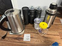 Front view of all items including kettle, water bottles, coffee grinder, glass mug, and thermos on wooden floor.