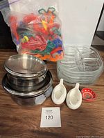 Full view of the lot showing stainless steel bowls with lids stacked, glass divided dishes stacked, ceramic sauce dishes, small decorative dish, and bag of plastic cookie cutters.