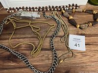 Five necklaces laid out on a wooden surface including two chain metal necklaces in gold and silver tones, a fine gold-tone chain, a wooden bead necklace with various shaped beads, and a dark brown bead necklace.