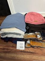 Photo showing folded sweaters or tops in light blue, white, gray, and dark colors. Pink crochet knit hat placed on top of dark garment and two pairs of transparent amber colored sandals with heart-shaped decorations on straps.