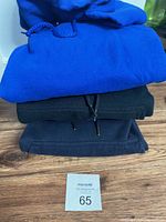 Stack of three folded men's hoodies or sweaters on wooden surface with a lot number card 65 visible