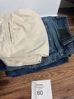 Stack of four pairs of jeans and pants with visible fabric textures and colors on wooden floor.