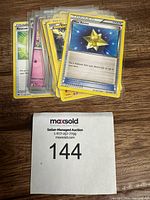 Photo showing a stack of assorted Pokemon cards including Hex Revive and Rescue Energy cards, placed on a wooden surface with a MaxSold tag.