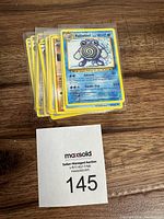 Stack of 11 Pokémon cards with Poliwhirl card on top, placed on a wooden surface with a MaxSold lot card beneath showing number 145