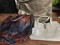Three purses/handbags grouped together: a metallic silver handbag, a dark blue and burnt orange large bag, and a white faux crocodile textured handbag.