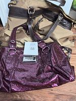 Lot contains two purses/handbags. The Guess handbag is shiny burgundy with embossed GG pattern and metal Guess plaque on front. The second handbag is tan with dark brown handles.