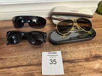 Four pairs of sunglasses photographed on a wooden floor including a beige Vogue case and black Tulot case