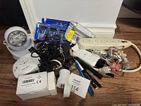 Full view of all miscellaneous household items arranged on wooden floor including LED light, power strips, cords, First Alert smoke detector, pens and tools.