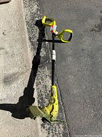 Full view of the Ryobi cordless string trimmer lying on asphalt and concrete surface, showing the handle, shaft and trimmer head with protective guard.
