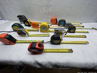 Overview of 14 assorted measuring tapes laid out on a white surface, showing extended metal tapes and varied casing colors and sizes.