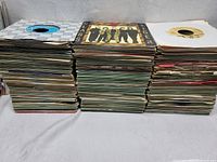 Stacks of over 300 45 RPM vinyl records piled horizontally in several stacks showing a variety of sleeves with different colors and designs.