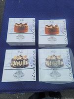 Four boxed new Cindeware Chic large square glass cake plates arranged in two rows of two, showing packaging with cake images on top.
