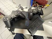 PlayStation 2 console with two black Sony controllers and a Sony remote on a table.
