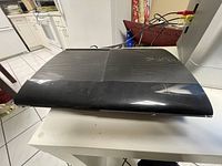 Photo of black Sony PS3 slim console with visible surface wear placed on a white table.