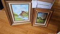 Two framed landscape paintings placed on hardwood floor, one larger with barn scene, one smaller with church scene