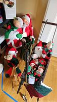 Photo shows large Santa and Mrs. Claus dolls standing side by side, several Christmas elves, a wicker sled filled with elf dolls, and a plush frog wearing a Christmas sweater.