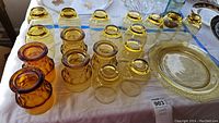Top view of vintage colored glass drinking glasses and decorative plate laid out on a tablecloth, showing mostly amber and yellow tinted glasses and one amber plate.