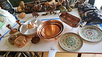 Overview of kitchen and decorative items arranged on table including copper bowls, candle holders, ceramic plates, copper containers and leaf-shaped dish