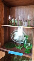 Wide shot showing items arranged on wooden shelf: 4 etched footed wine glasses in green, salt and pepper shakers (green and clear), a small footed dessert dish, a footed candle holder, round plate, and a large platter at the back.