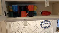 Fiesta Ware mugs stacked and arranged in varying colors of blue, yellow, black, orange, red, and green alongside a red lidded chili pepper dish with black spoon on a shelf.