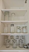 Glass jars on white shelves, various sizes with metal bale wire lids and some with zinc screw lids.